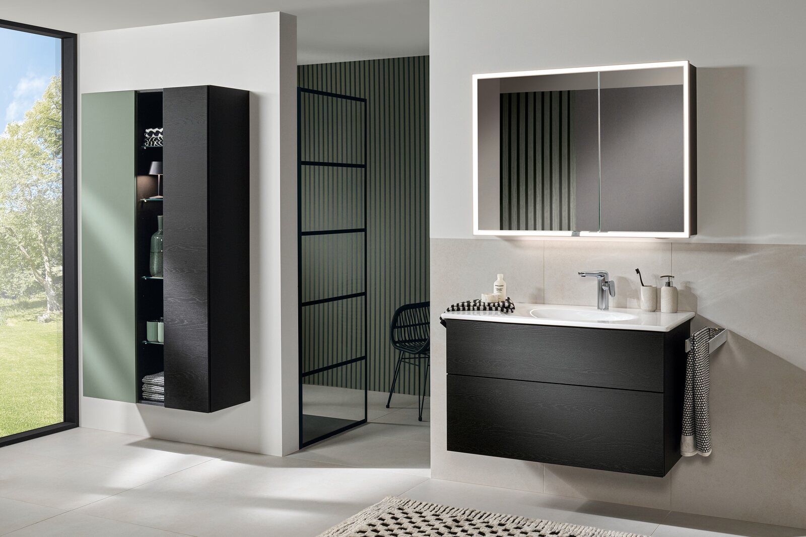 Badezimmer mit schwarzem Waschtisch-Unterschrank mit zwei Auszügen und weißem Waschtisch unter einem beleuchteten Spiegelschrank, links ein schwarzer Hochschrank mit offenem Regalfach neben einem bodentiefen Fenster